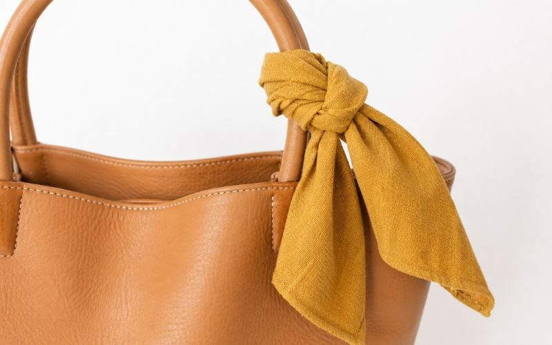 Mustard yellow cotton scarf tied in a classic knot around a tan leather tote bag handle, overhead close-up